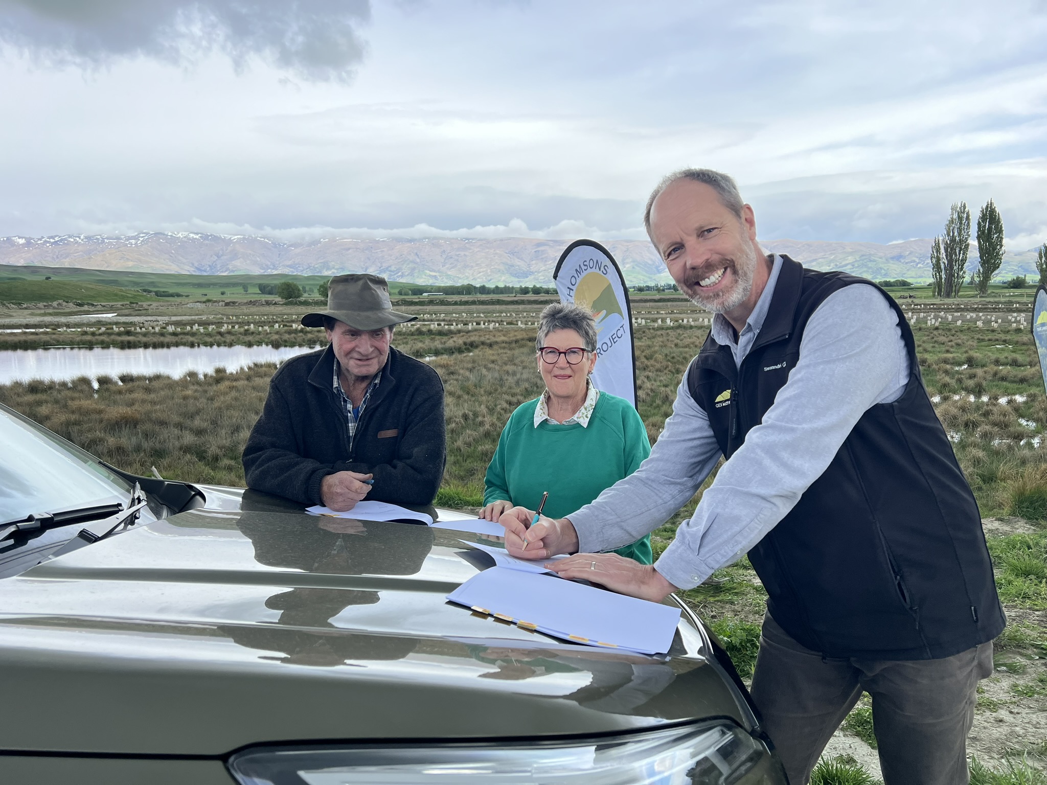 Peter and Robin McLeod stand around a car with Dan Coup, who is about to sign papers on the car bonnet, wetland in the background